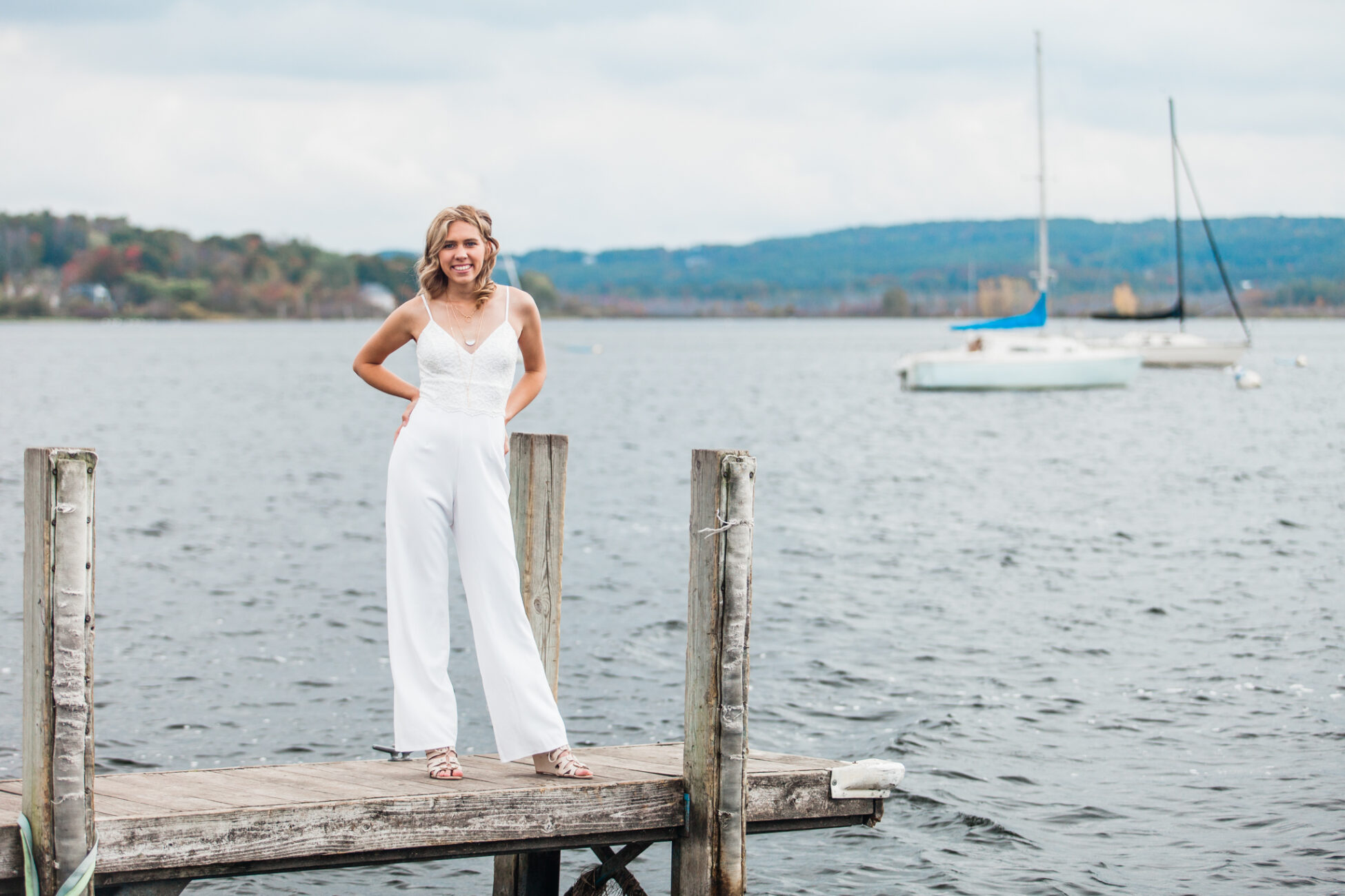 senior girl standing on a dock next to water wearing white jumpsuit with sailboats behind